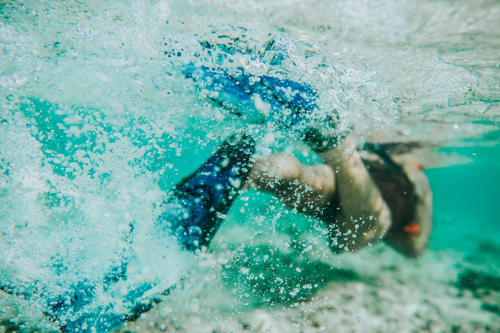 Underwater, a swimmer in fins dives into the turquoise water among the bubbles.