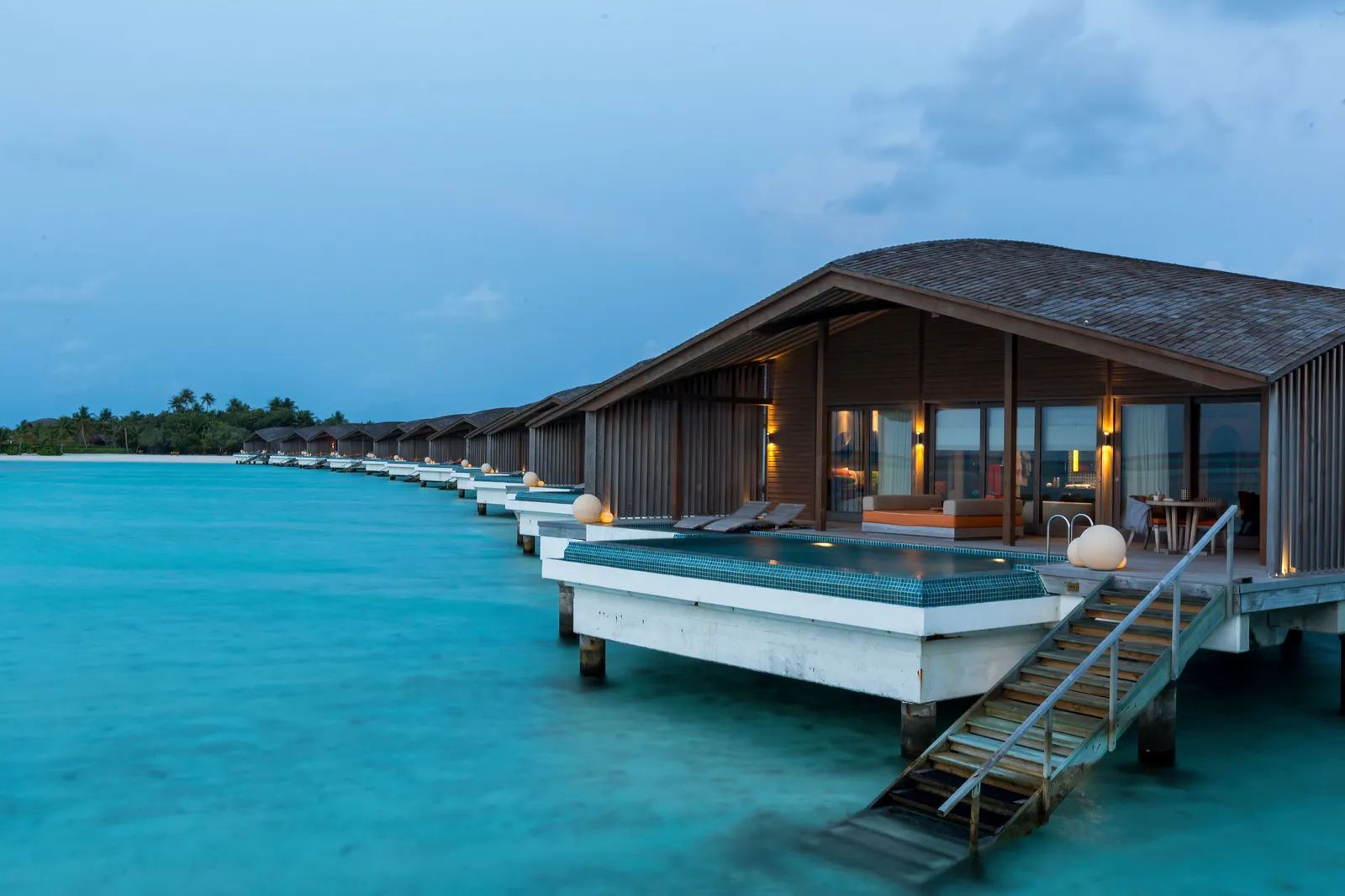 The water villas at dusk, lights on, a pool on the deck and the steps going down into the lagoon.