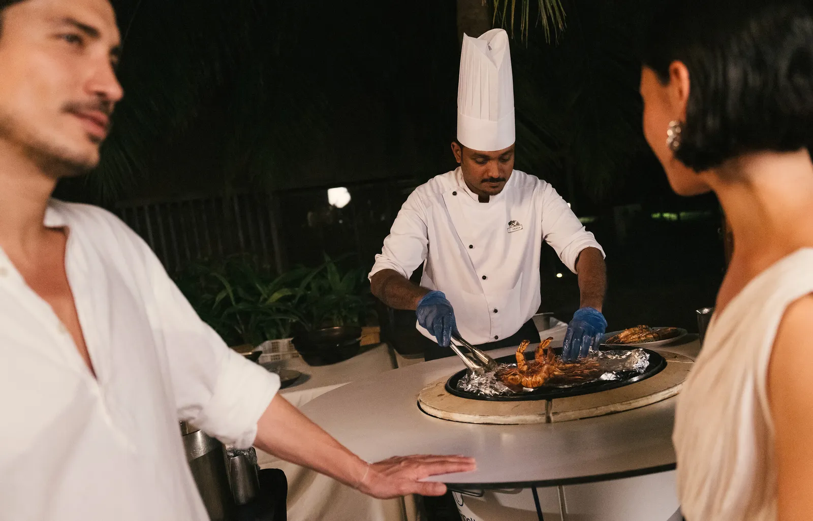 A chef cooking at the table in front of a couple, one evening among the greenery and low lights.