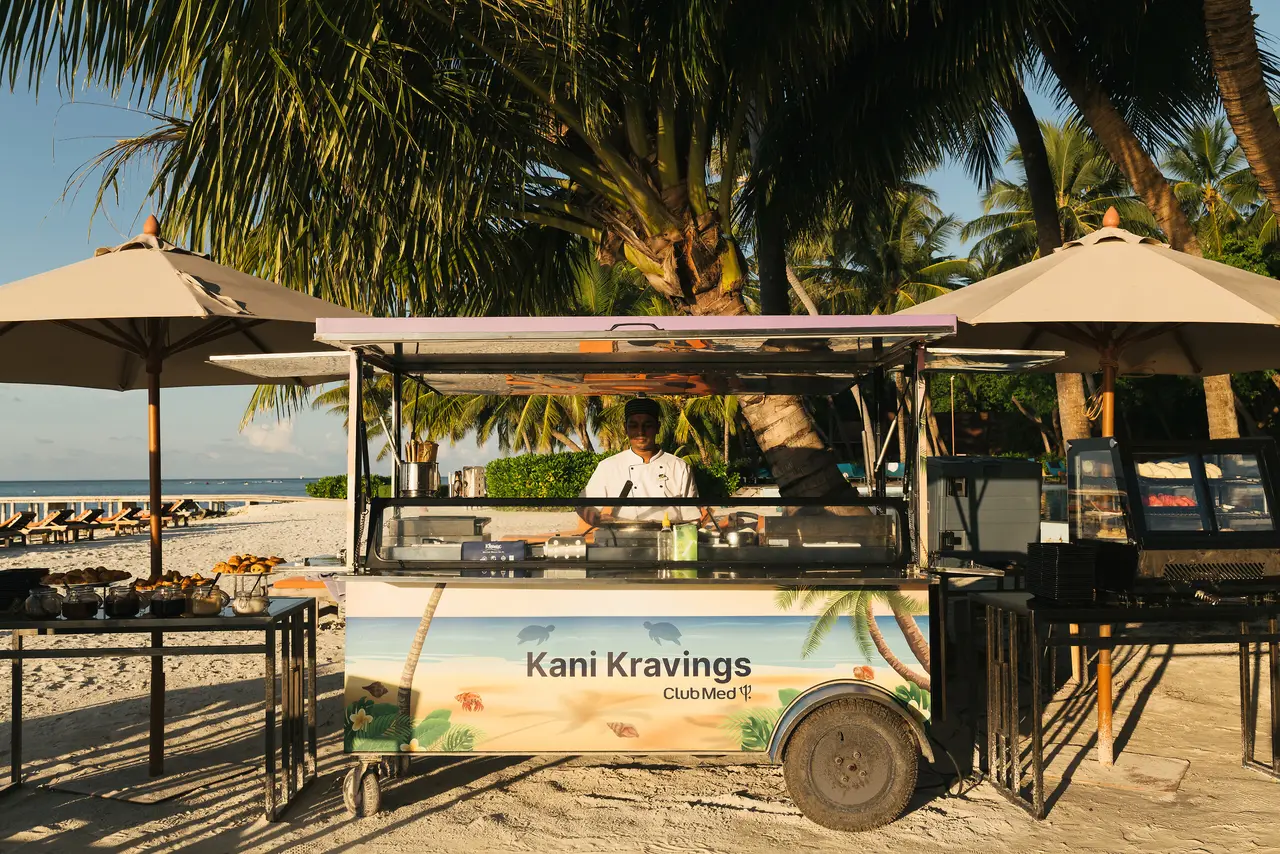 The Kani Kravings food truck on the beach, under the parasols, with a cook at the counter.