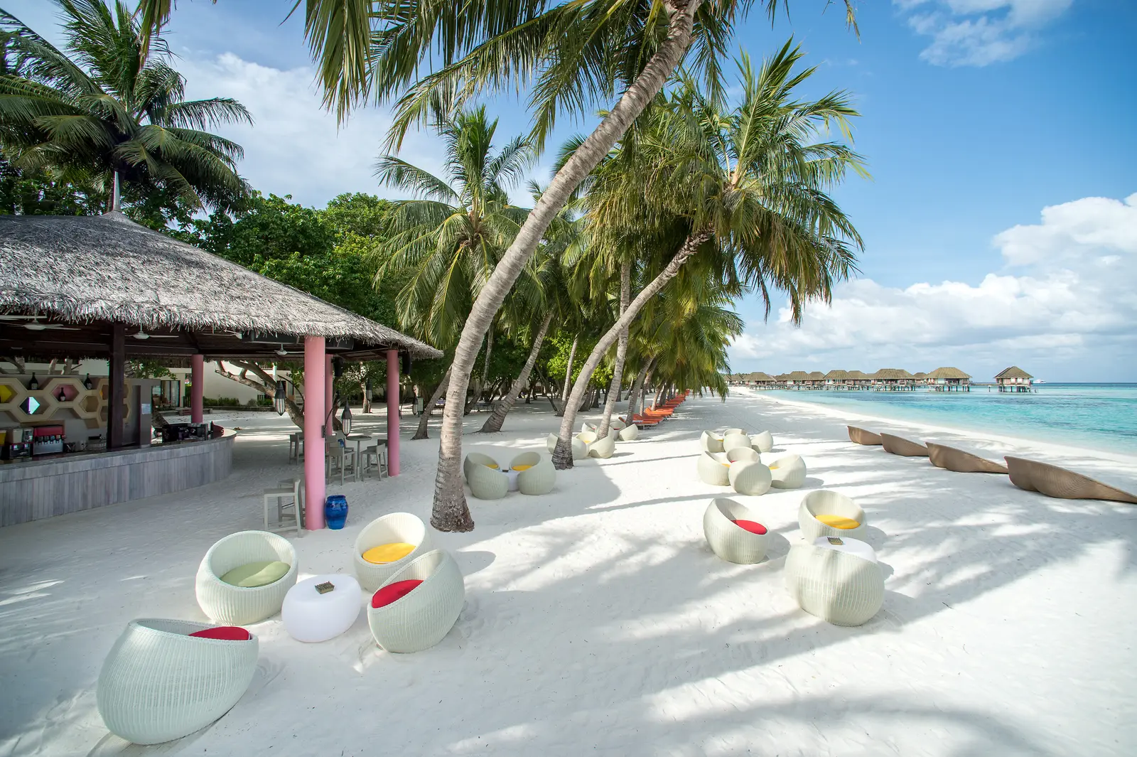 The Sunset bar on the beach, its thatched roof and white seats on the sand among the palms, the overwater suites in the distance.