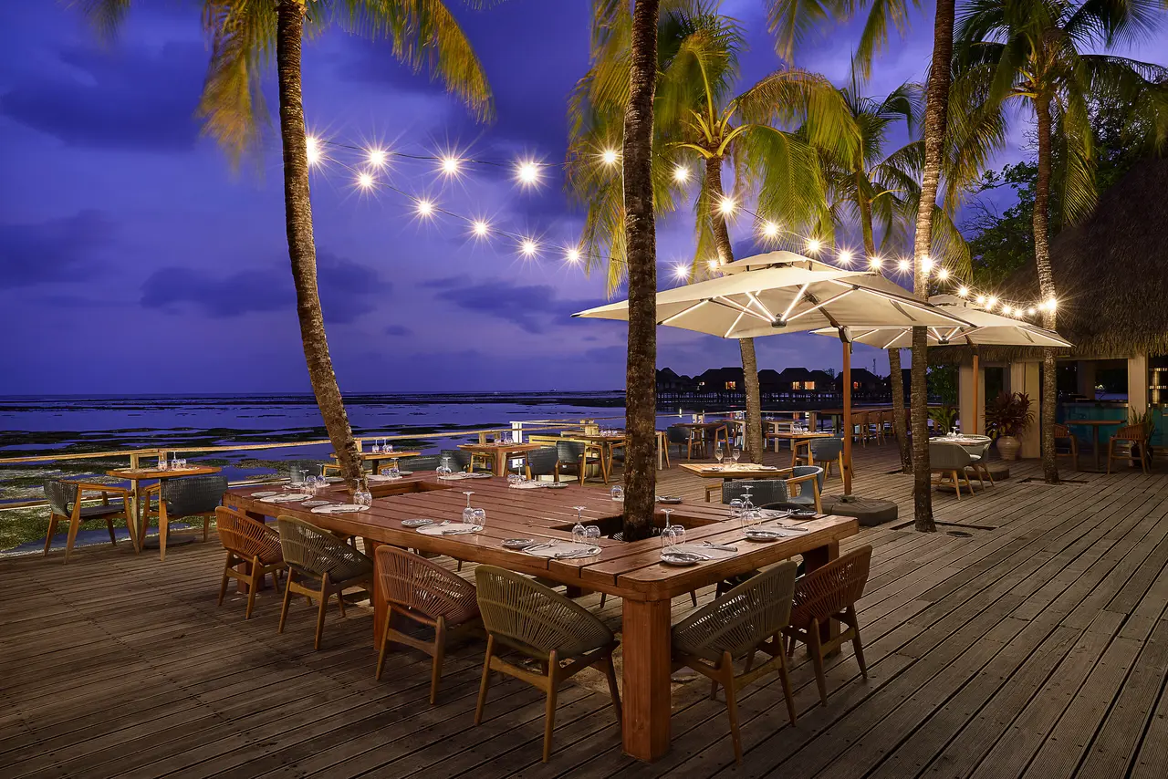 The Kandu’s lagoon deck at night, with its long wooden table, the parasols and the string lights among the palms.