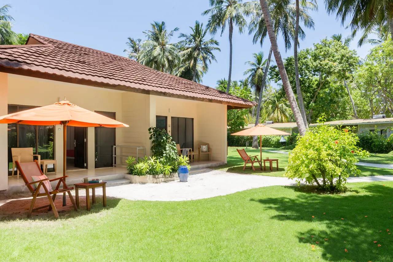 A single-storey garden bungalow, its tiled roof, loungers under an orange parasol and the lawn among the palms.