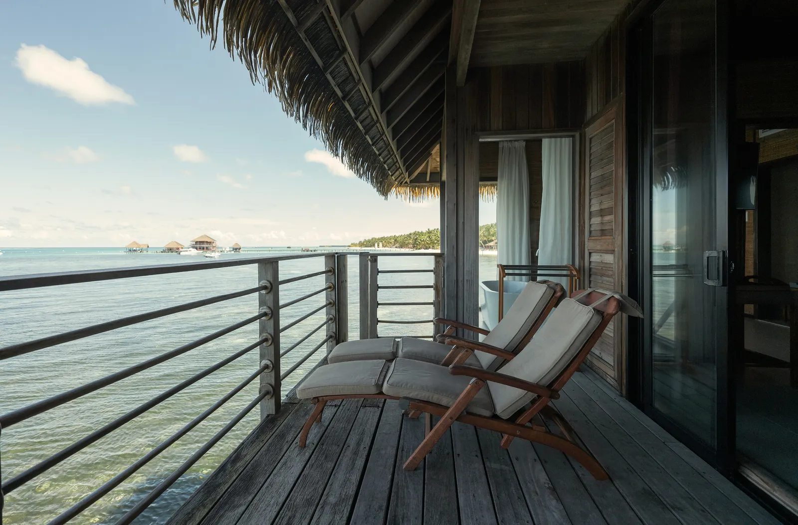 A panoramic-view overwater suite’s deck: 2 loungers under the thatch and the ocean ahead.