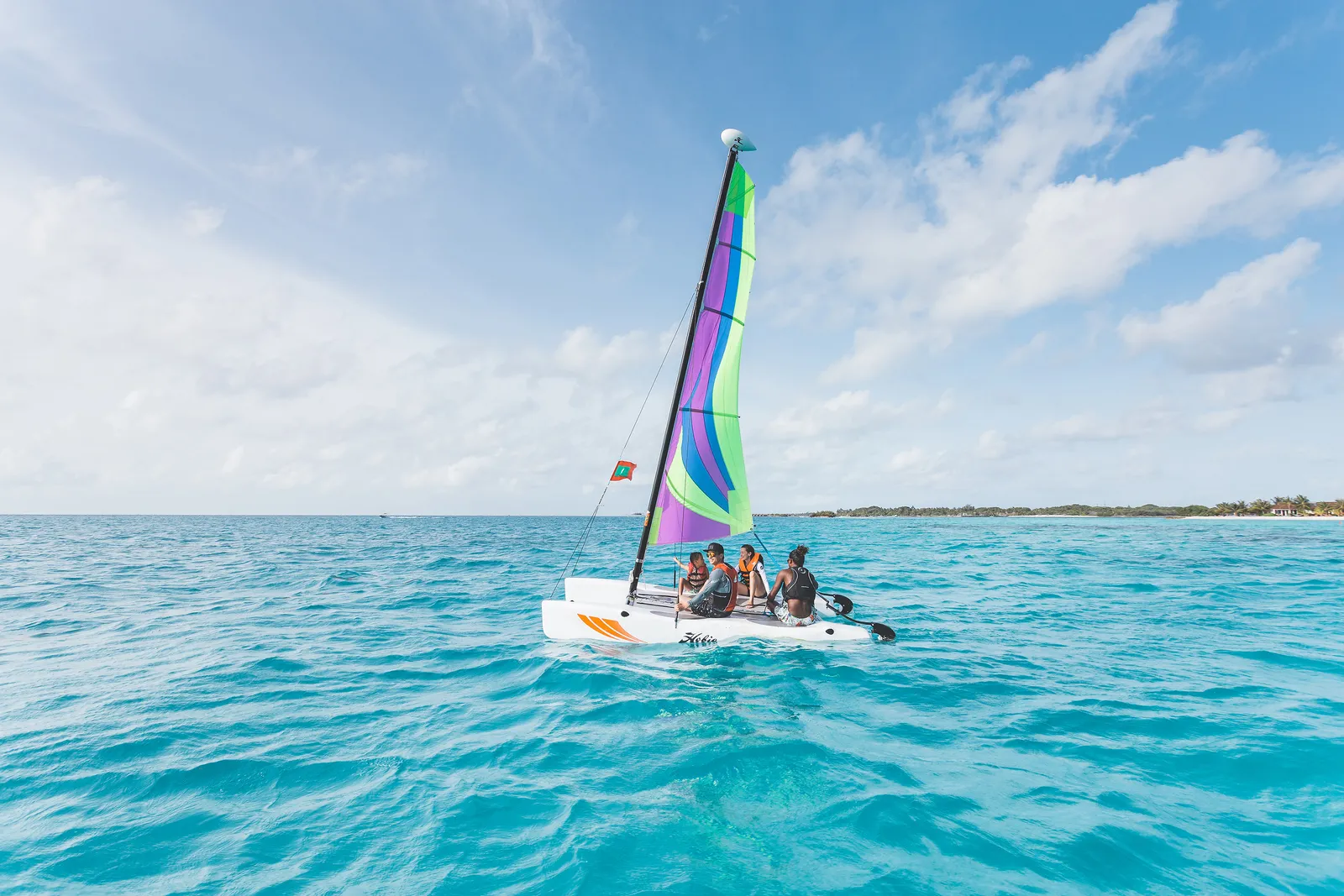 A small catamaran with a bright sail and 3 people aboard on the turquoise lagoon.