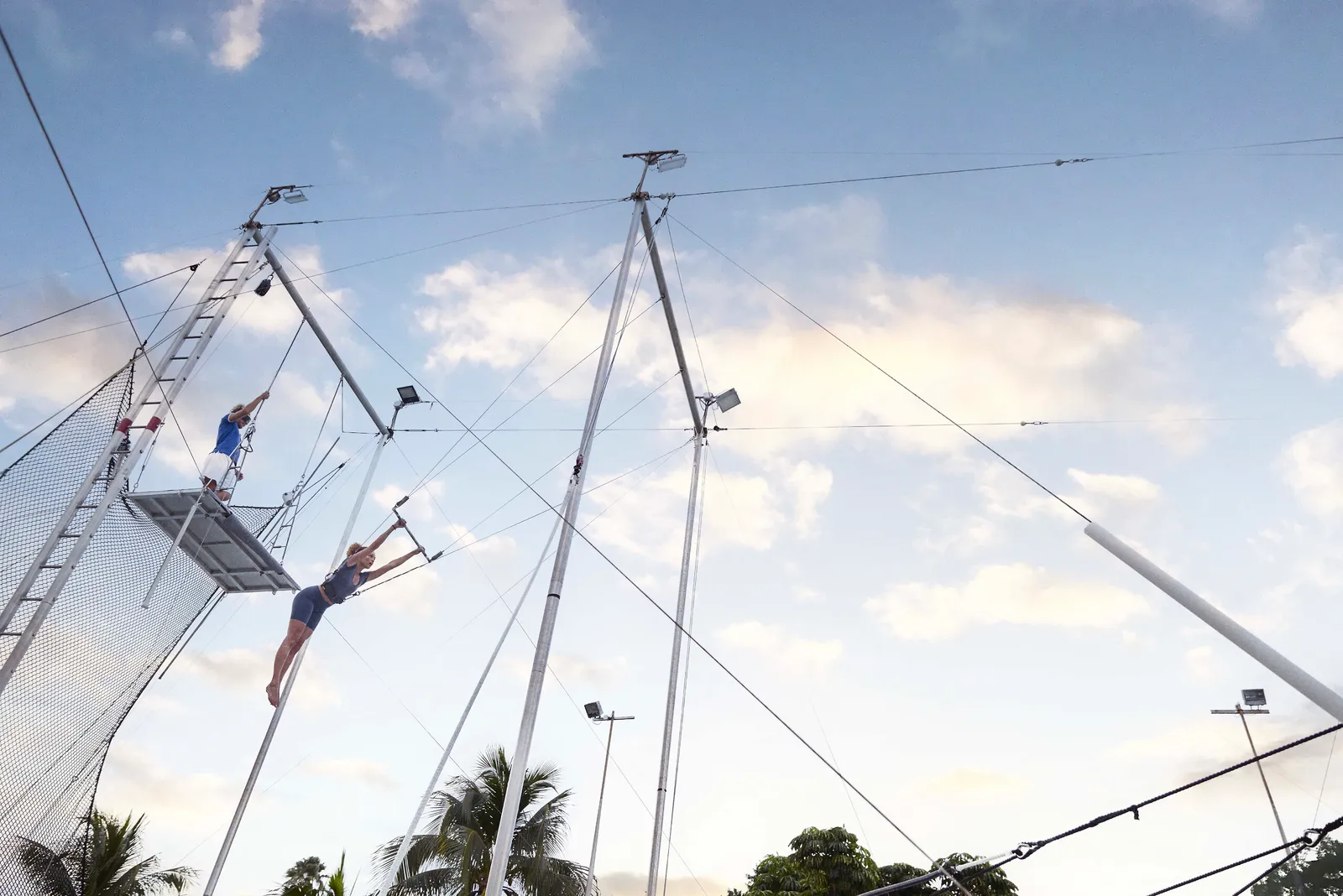 The flying trapeze on the beach: a flyer in mid-air between the platform and the bar, against the sky.