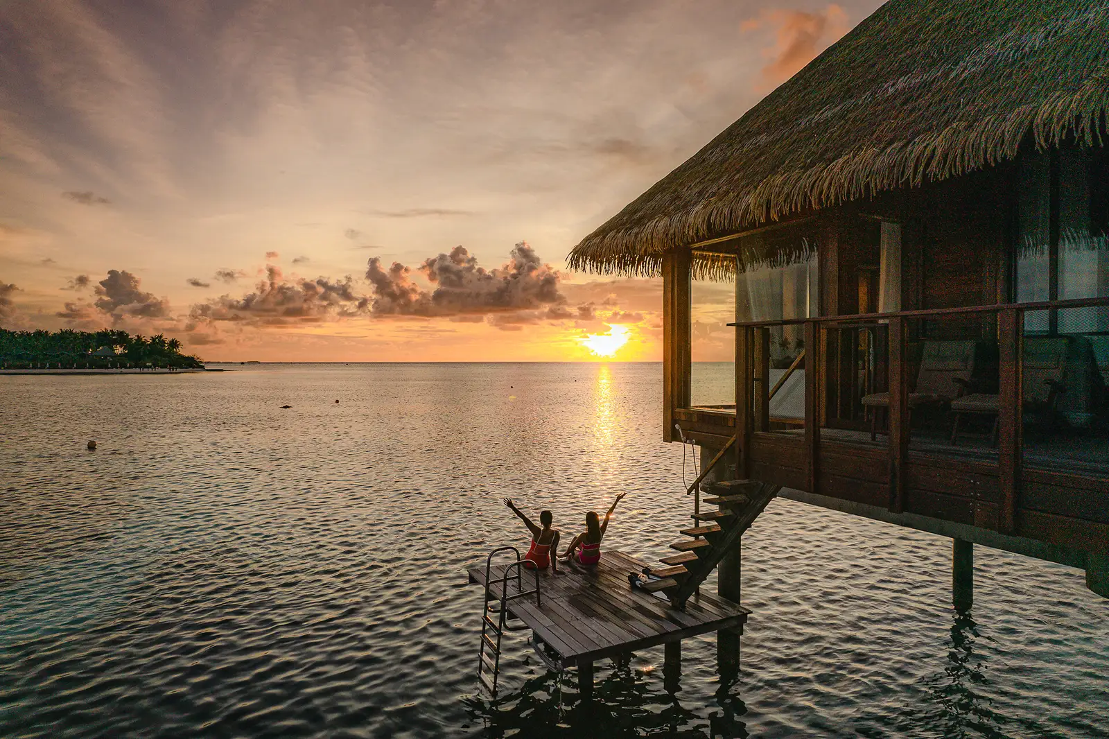 2 people with their arms raised on the steps of an overwater suite, at sunset over the lagoon.