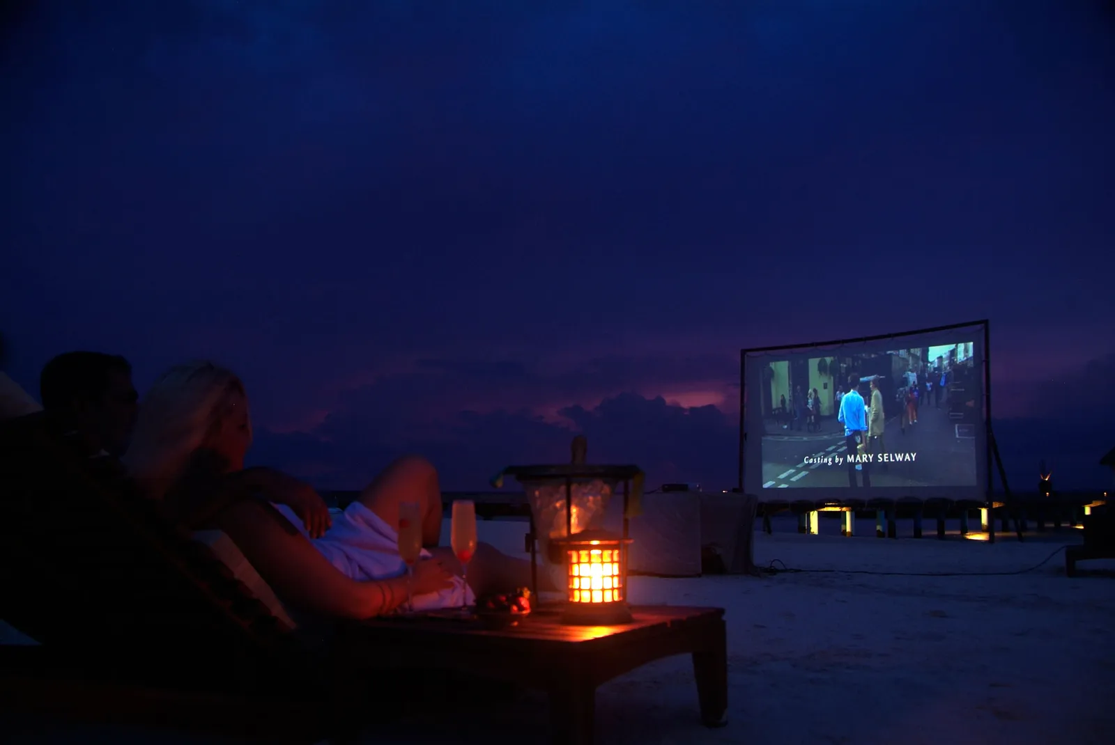 The beach cinema at night: the screen lit on the sand and guests on loungers among lanterns.