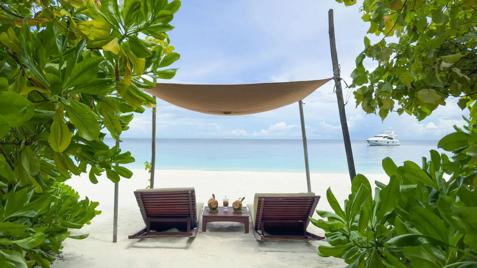 2 loungers under a shade sail on the beach, framed by bushes, with the sea and a boat beyond.