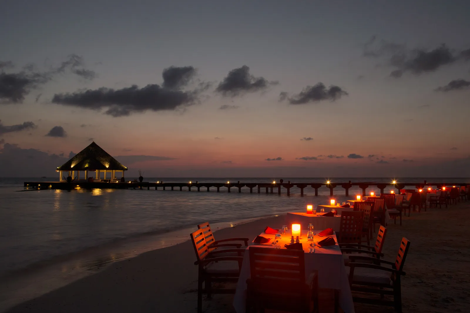 Tables laid on the sand at dusk with candles lit, and the pavilion at the end of the jetty glowing over the sea.