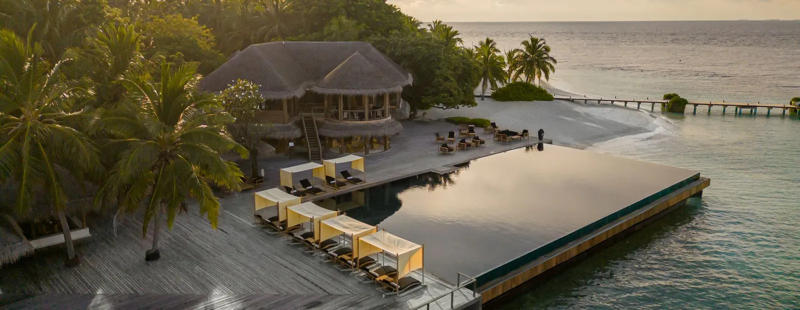 The main infinity pool at sunset, loungers under canopies, a thatched pavilion among the palms and the jetty over the sea.