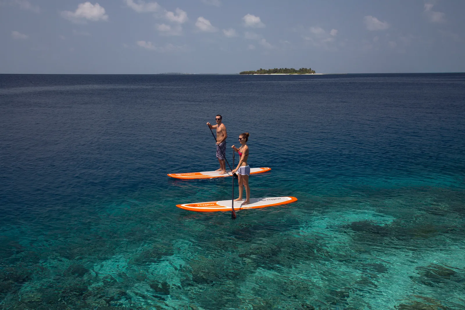 2 people stand-up paddling on the lagoon, seen from above, an island on the horizon.