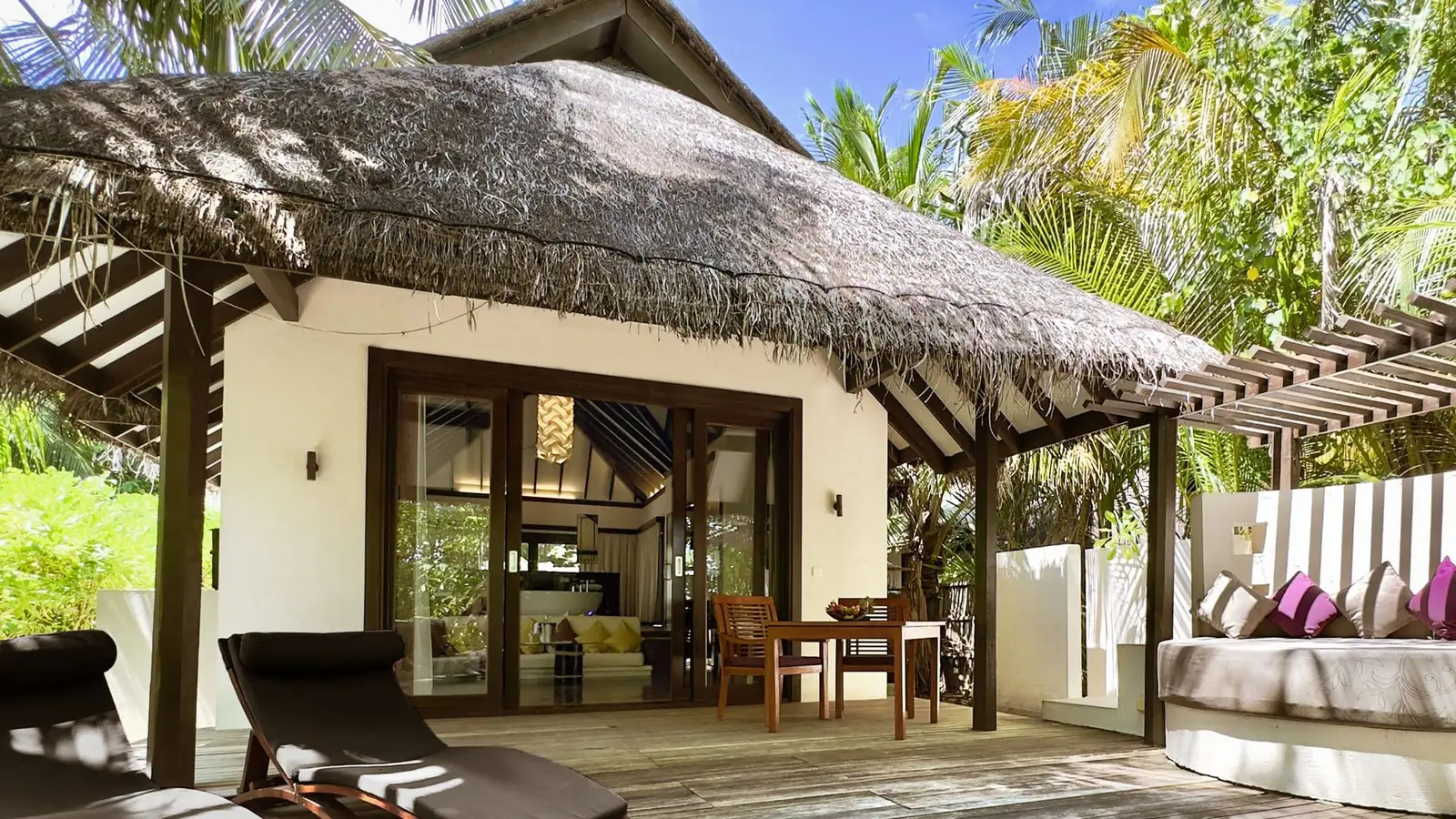 A Beach Villa with Pool’s deck: loungers on the timber, the thatched villa with its glass doors open and the outdoor sofa.