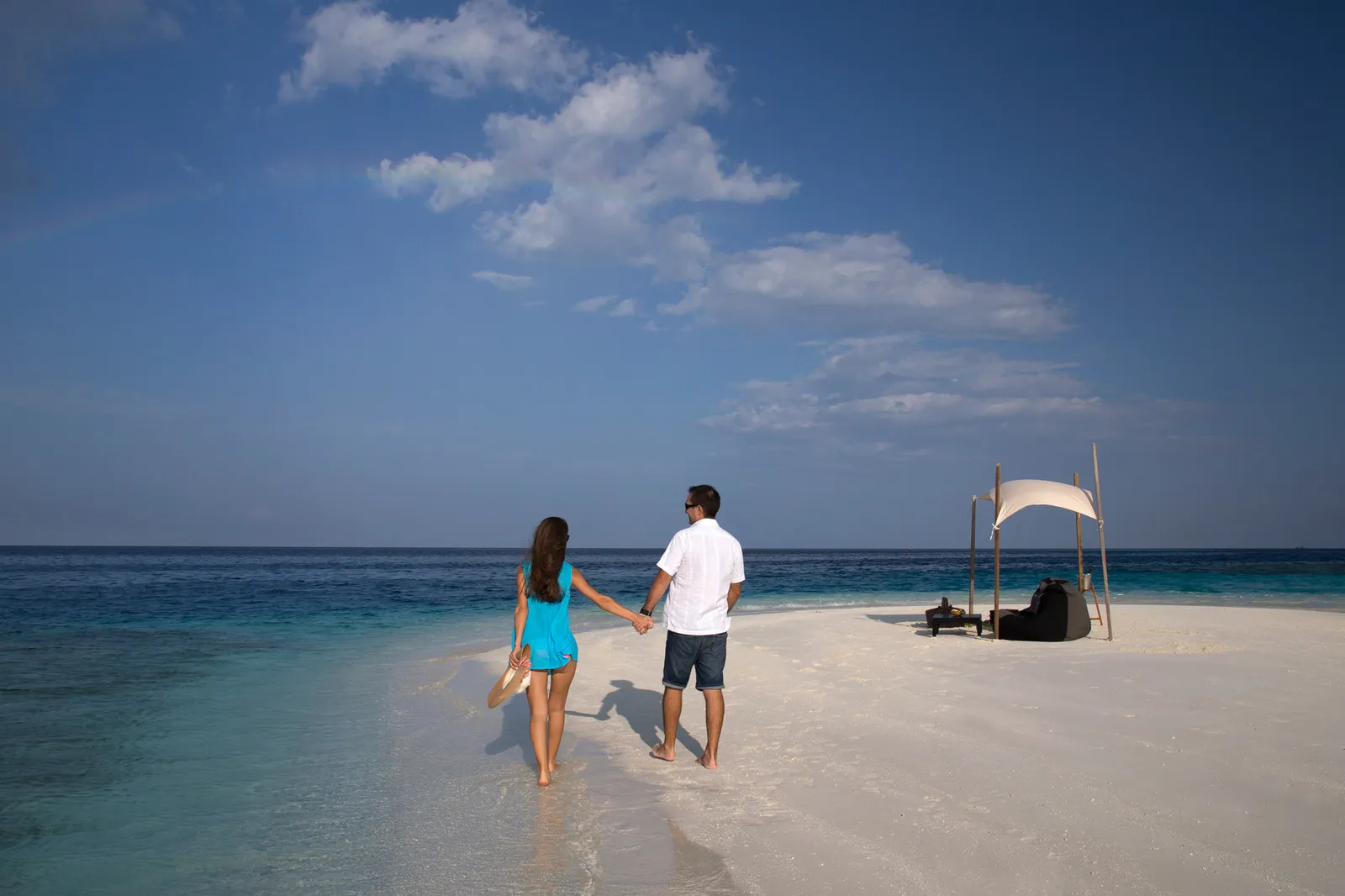 A couple walking hand in hand on a sandbank, a sunshade and picnic cushions behind them.