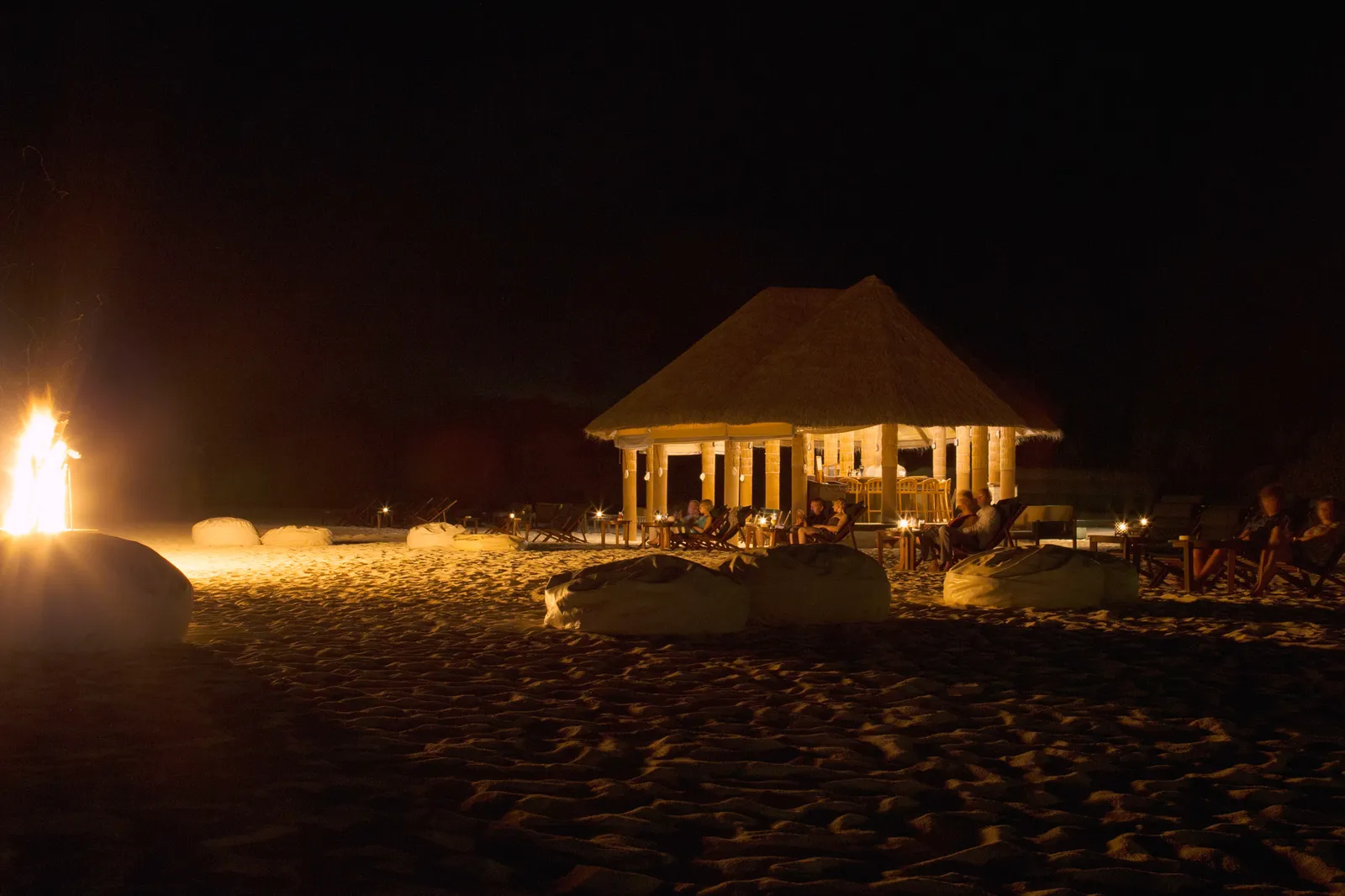 The Beach Bar’s pavilion lit up at night on the sand, with a fire burning.
