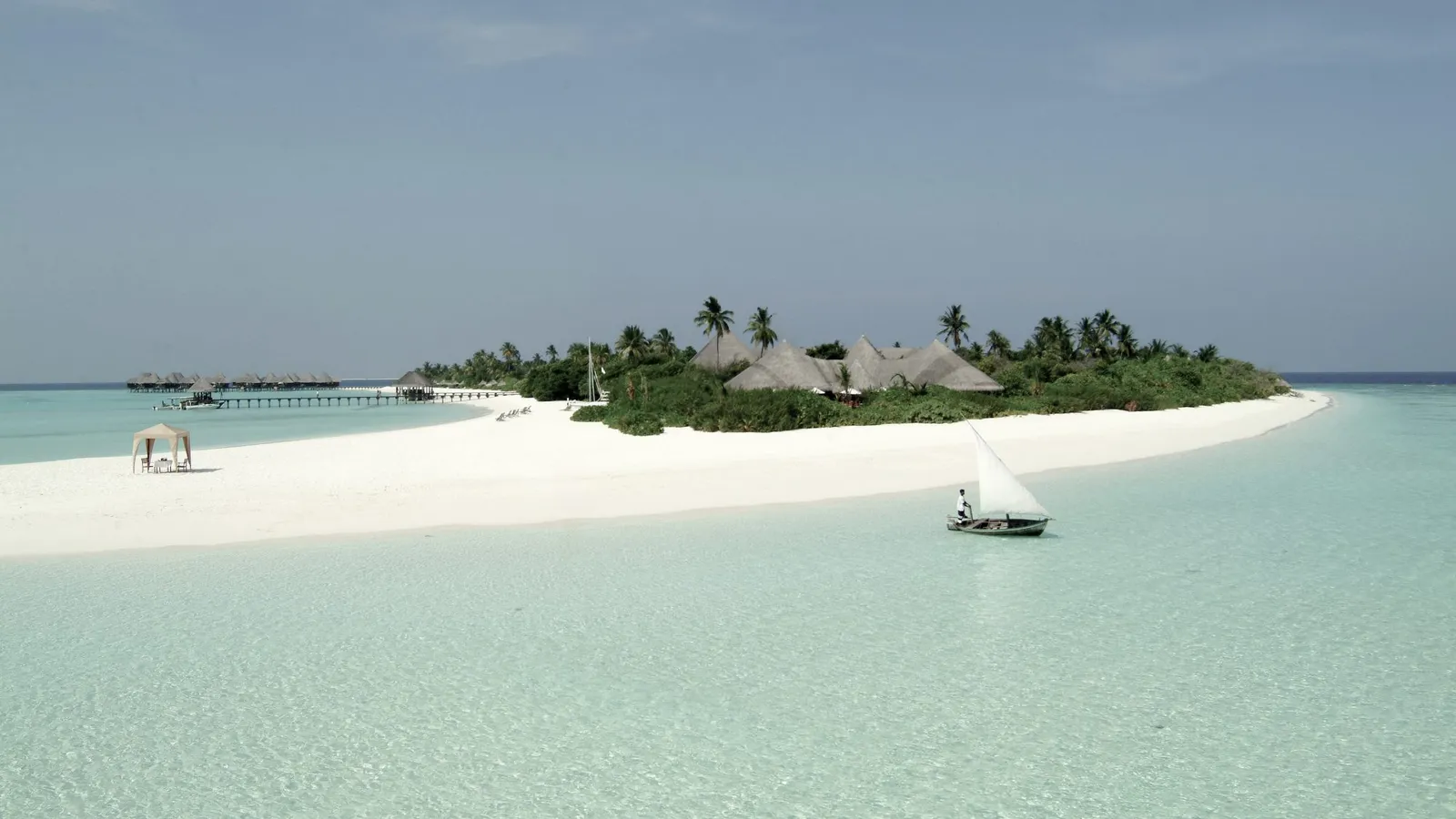 Dhuni Kolhu seen from the lagoon: the spit of white sand, the palms, the water villas beyond and a sailing boat.