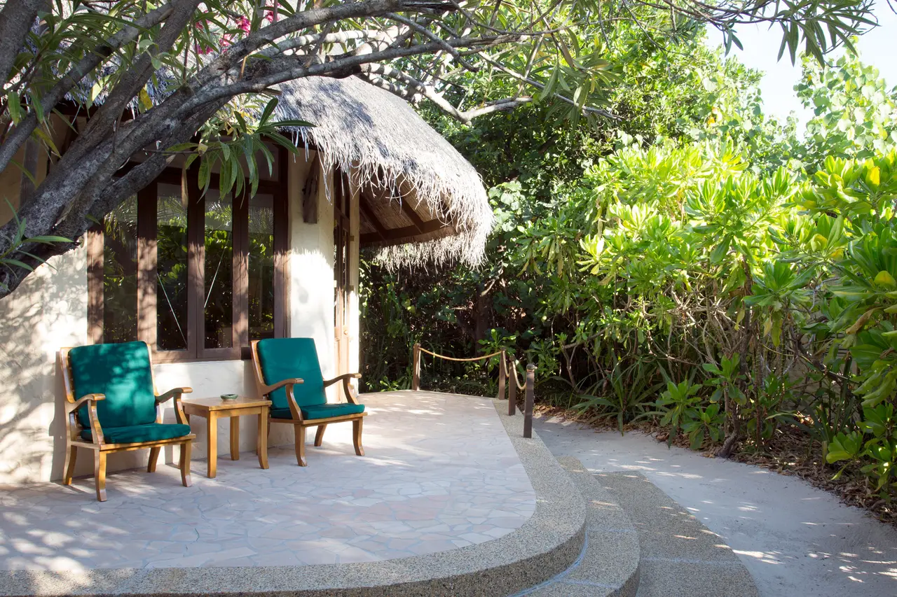 A Sunset Beach Villa’s terrace, 2 green armchairs and a side table in the shade of the thatch, among the plants.