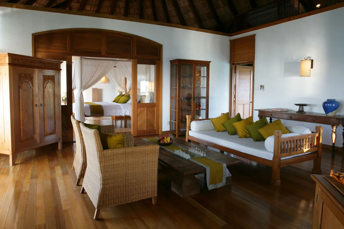 A Sunset Lagoon Villa’s living room, with timber floors, a sofa, carved cabinets and the bed beyond the arch.