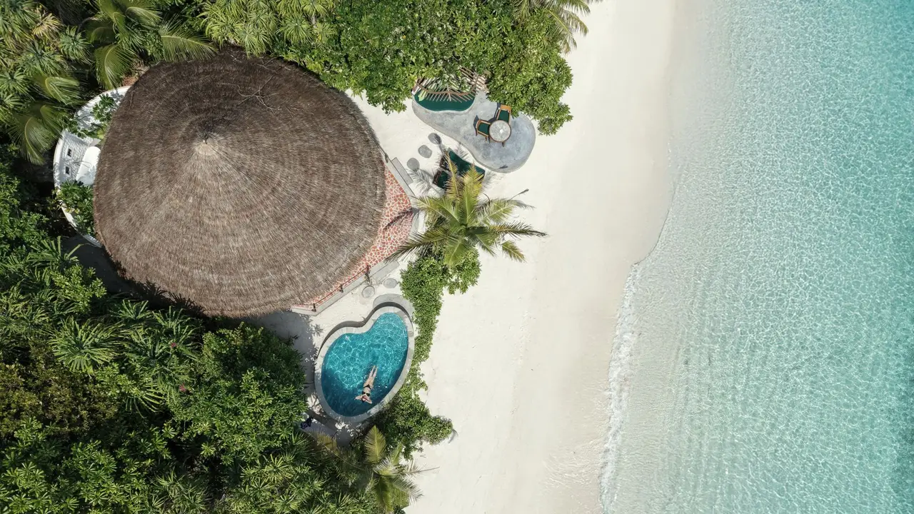 From above, a villa’s thatched roof among the trees, its pool and the white beach running into the lagoon.
