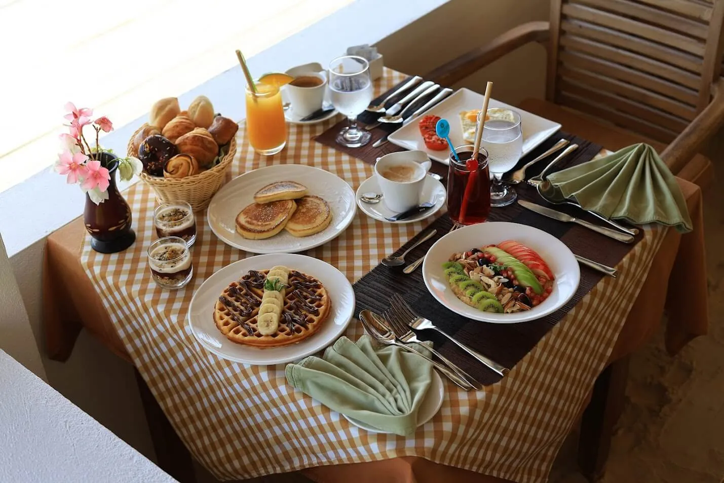 Breakfast at the table: pancakes, a waffle, fruit, croissants and juice on a checked cloth.
