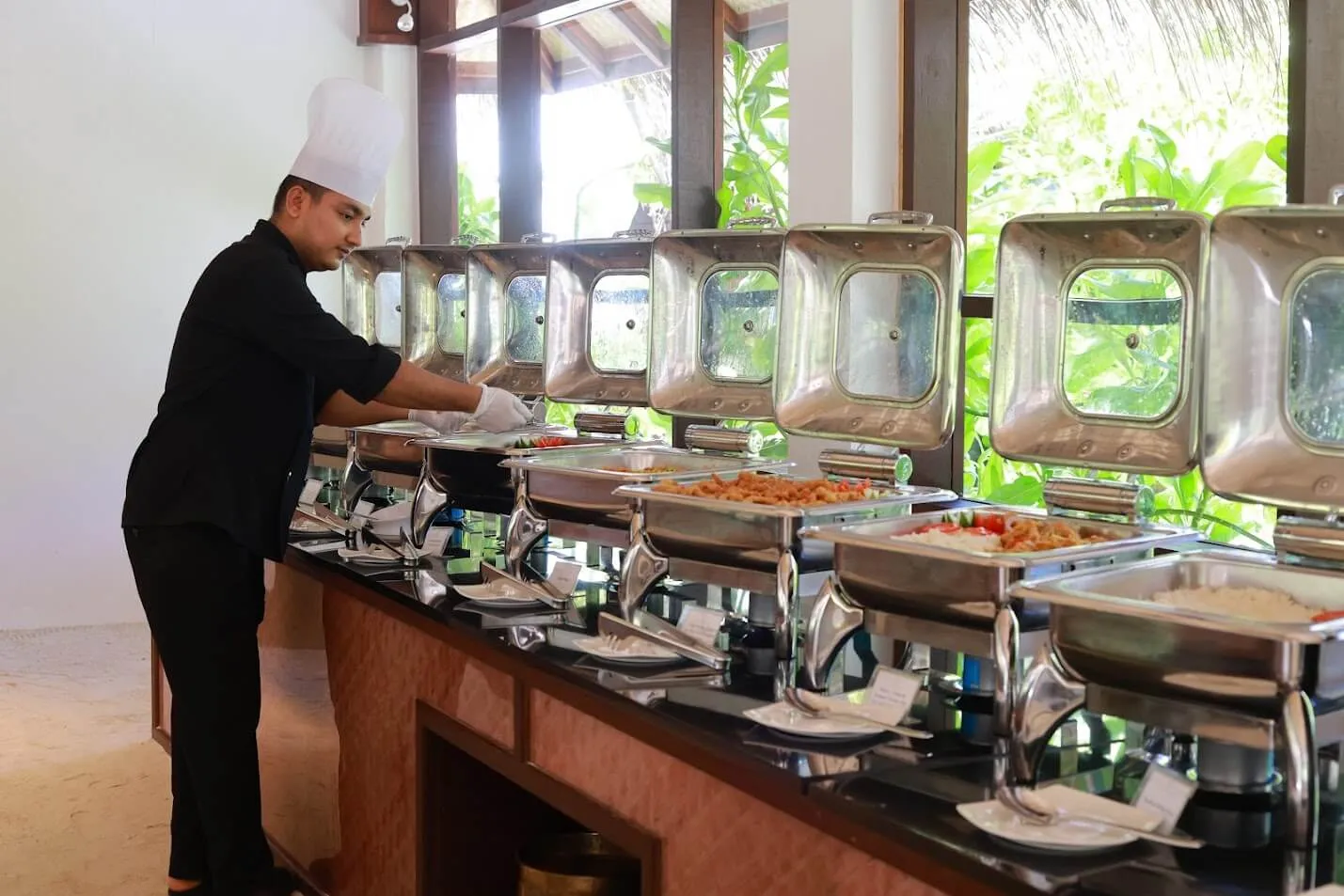 A chef at the buffet counter, among the steel chafing dishes, with greenery beyond the windows.