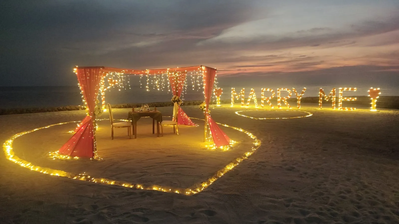 A proposal set up on the beach at dusk: a heart of lights on the sand, the table and the words Marry me.