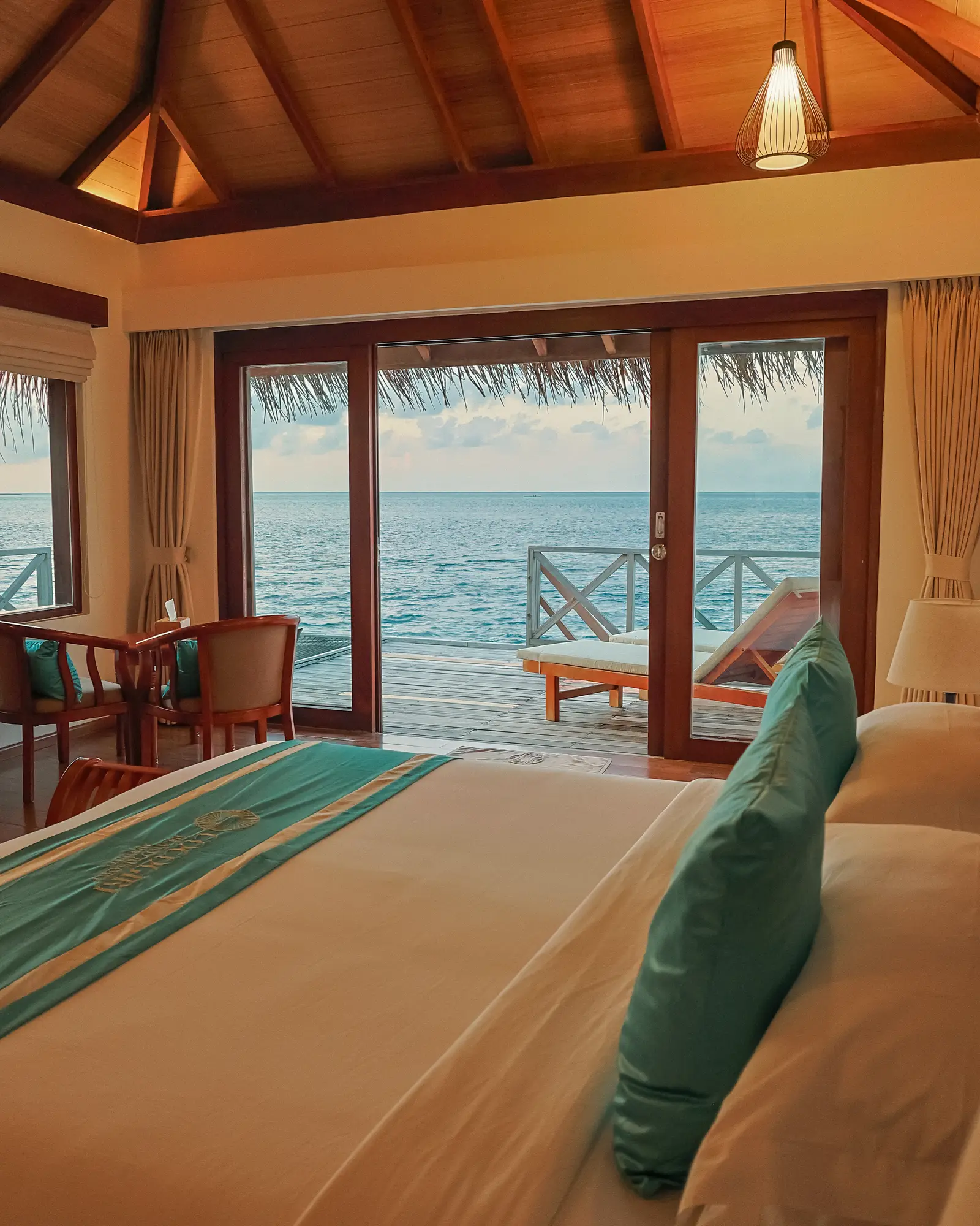 A Water Villa bedroom: the king bed with teal cushions and the glass doors open onto the deck and the sea.