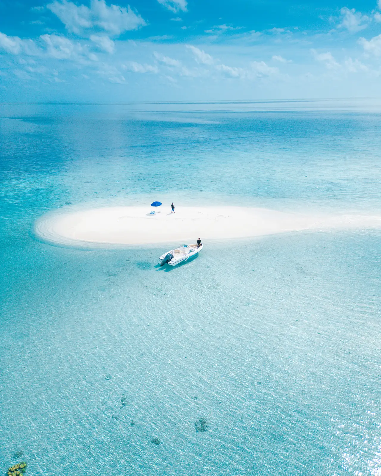 A sandbank in the middle of the lagoon seen from above, with 2 people, a parasol and the boat alongside.