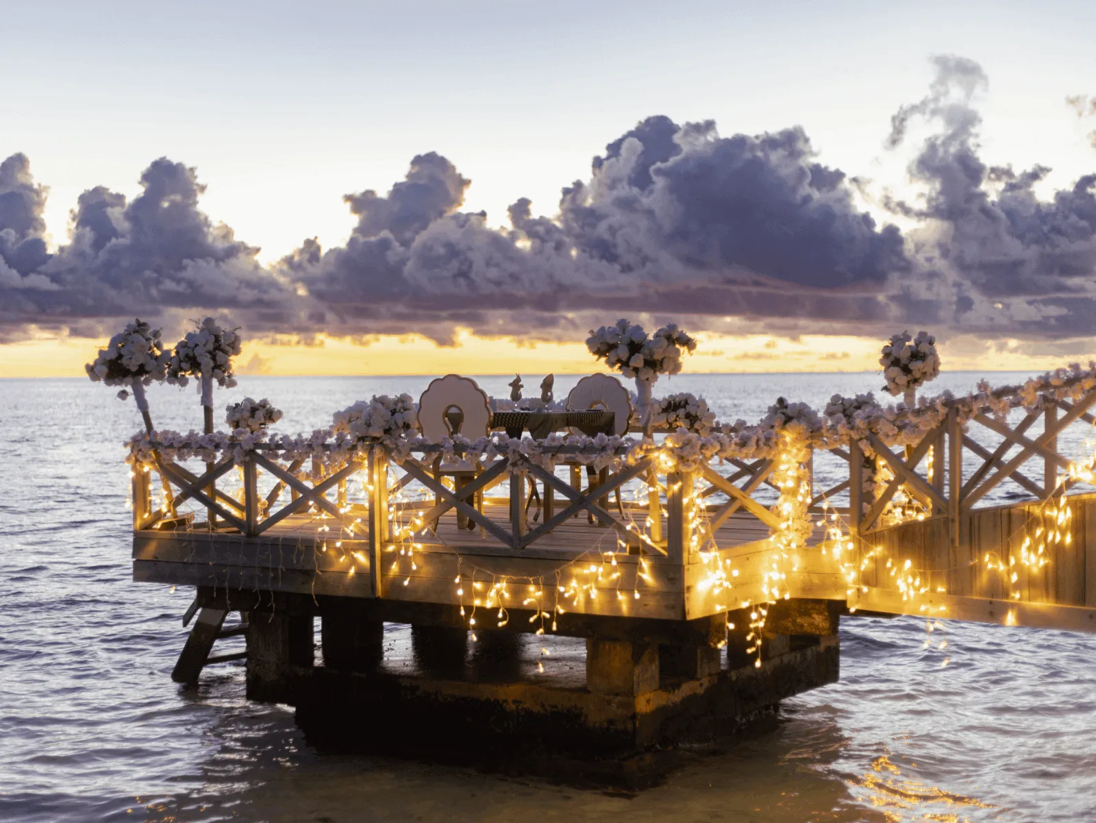 The Sunset Pavilion laid for dinner, its lights strung along the railing above the water, at sunset.