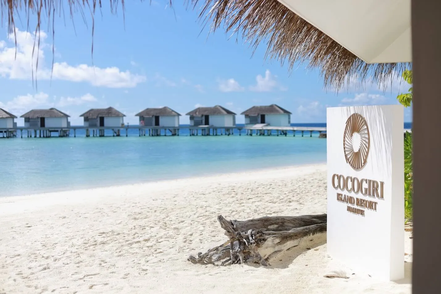 The Cocogiri sign on the beach, driftwood on the sand and the row of water villas across the lagoon.