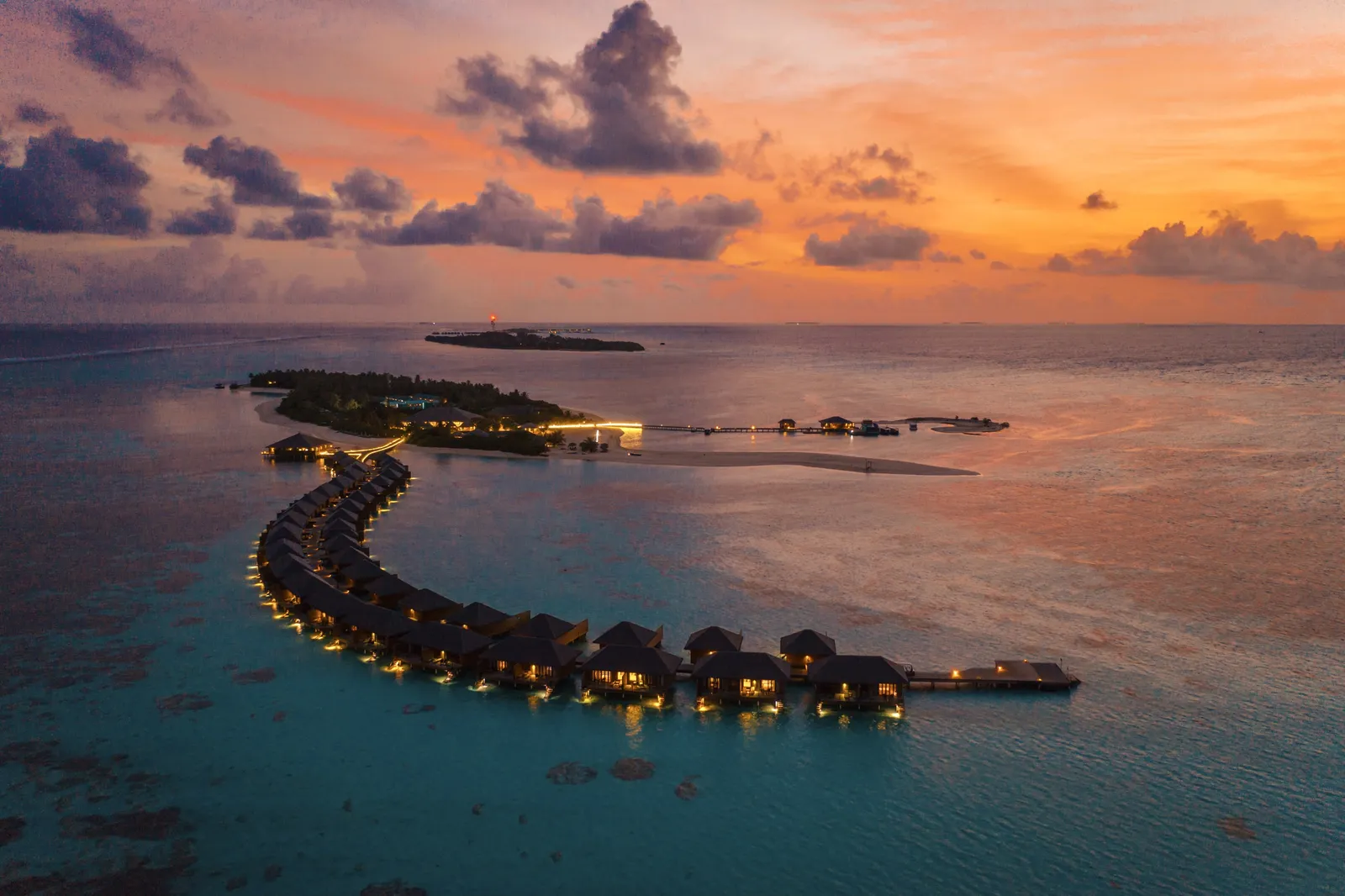 The arc of lagoon villas lit up at dusk, the island behind