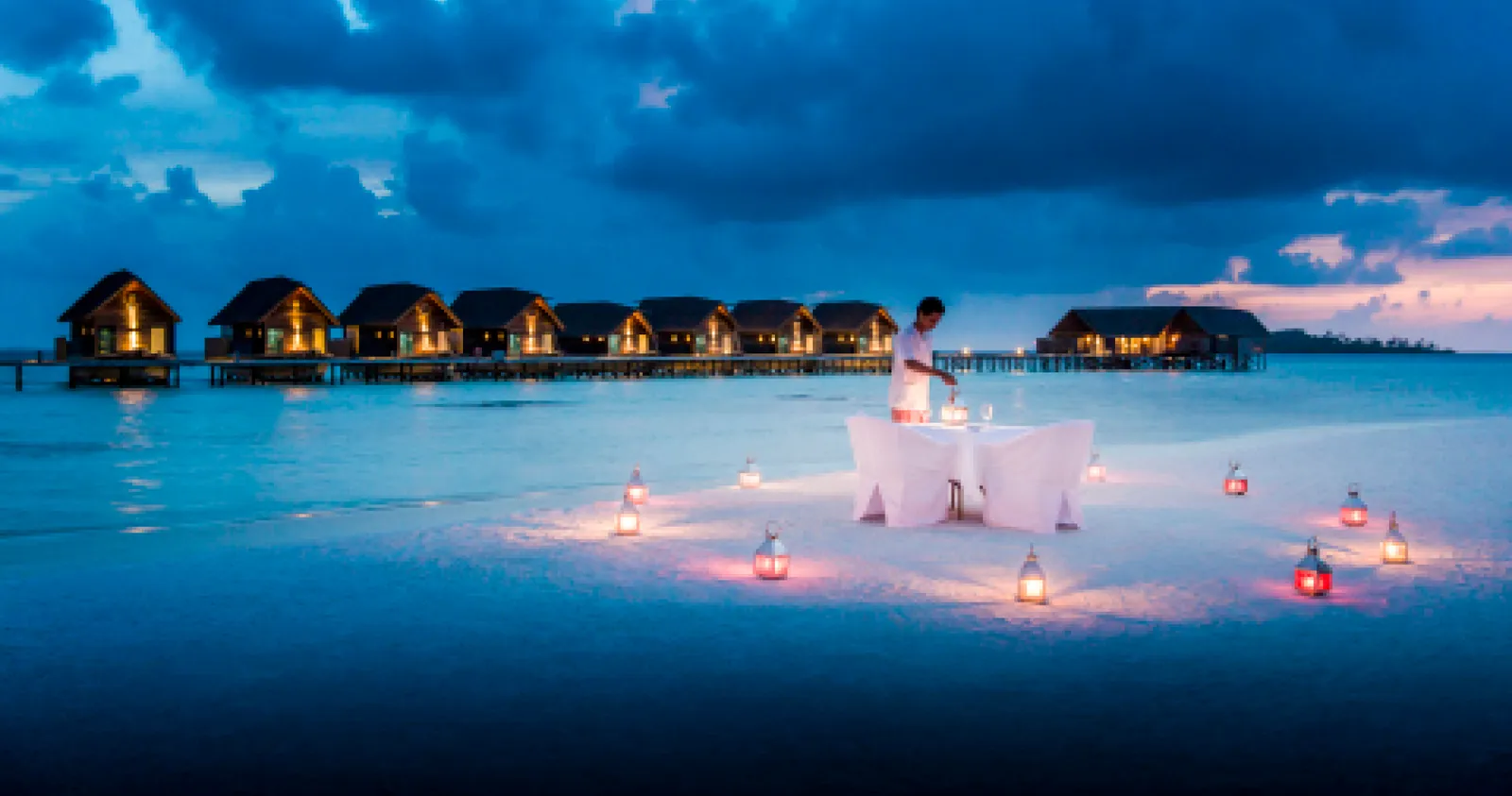 A private dinner in the shallow lagoon at dusk, among lanterns, with the overwater villas behind