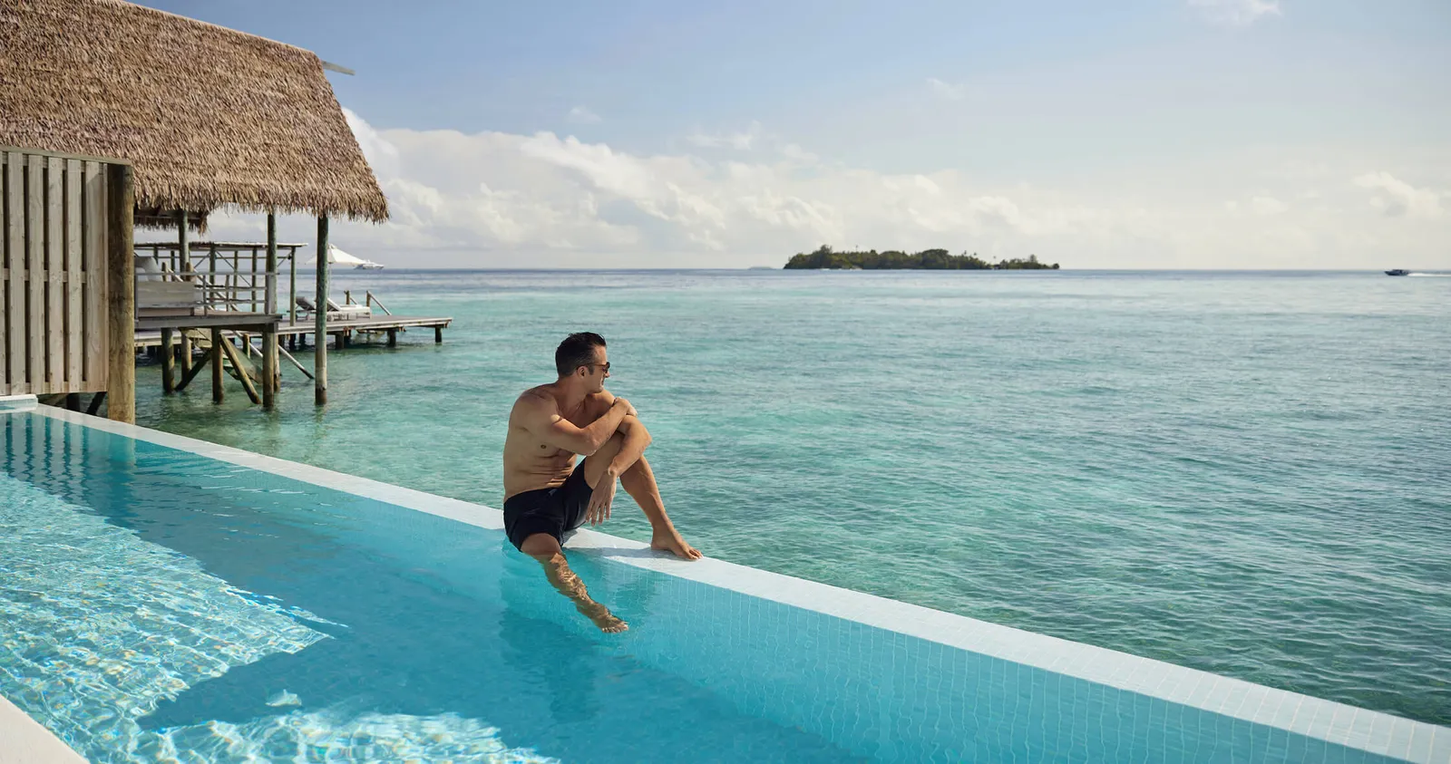 A guest sitting on the edge of his villa pool, facing the lagoon and a distant island