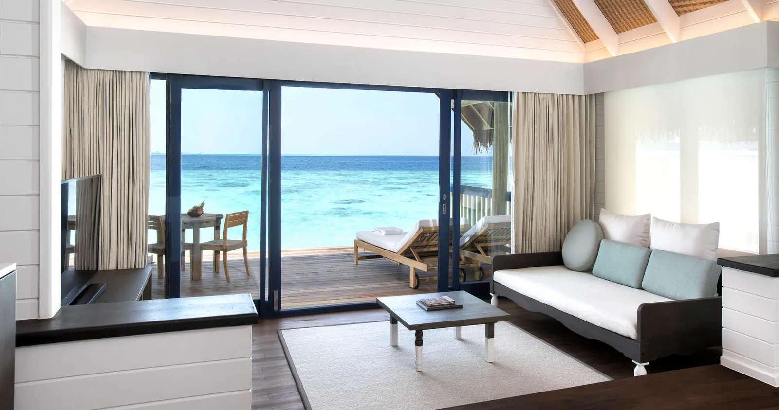 The living room of a Dhoni Water Villa, glass doors open to the deck and the lagoon