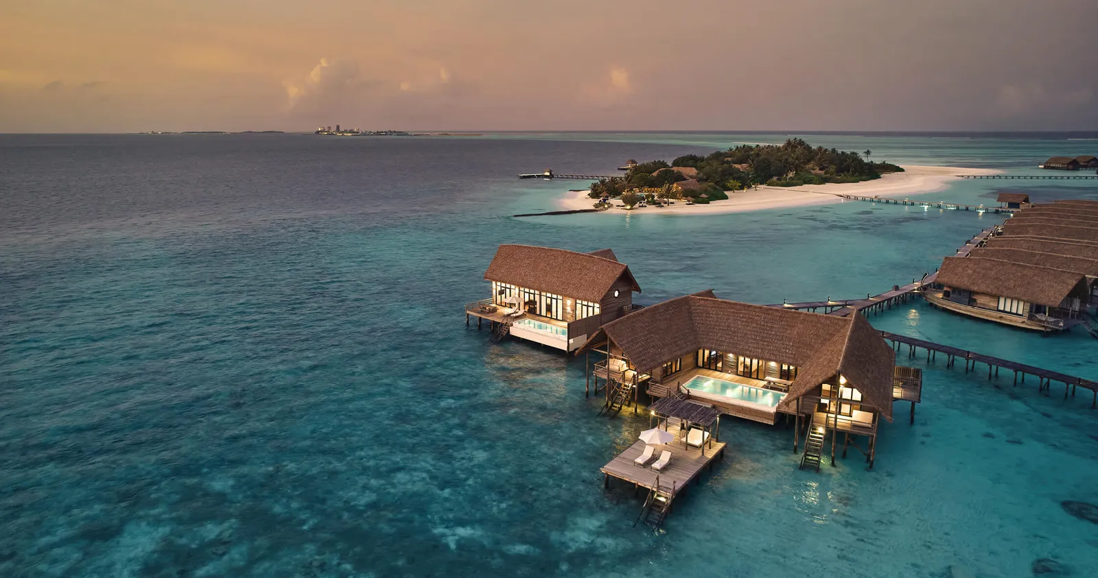 The large overwater pool villas at sunset, seen from above, with the island behind