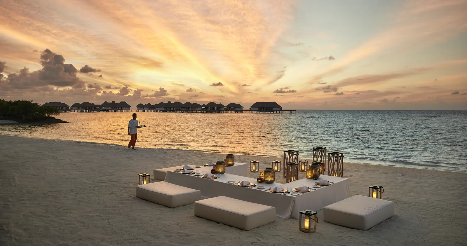 A dinner laid on the beach at sunset, with lanterns and white cushions, and the water villas on the horizon.