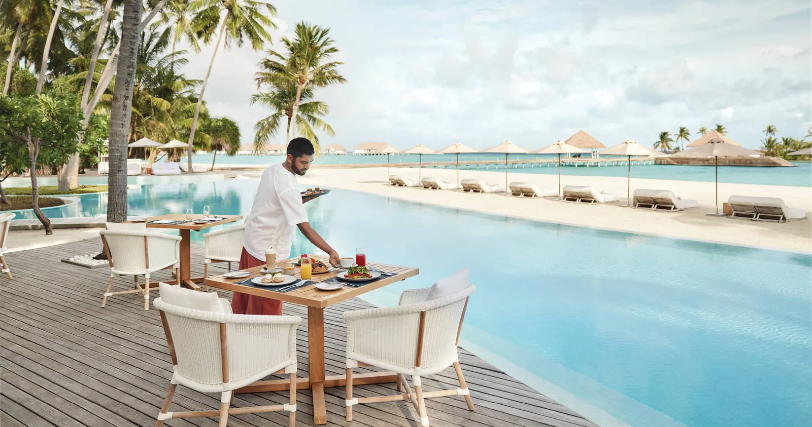 A waiter laying a table beside the pool, with parasols on the beach behind.