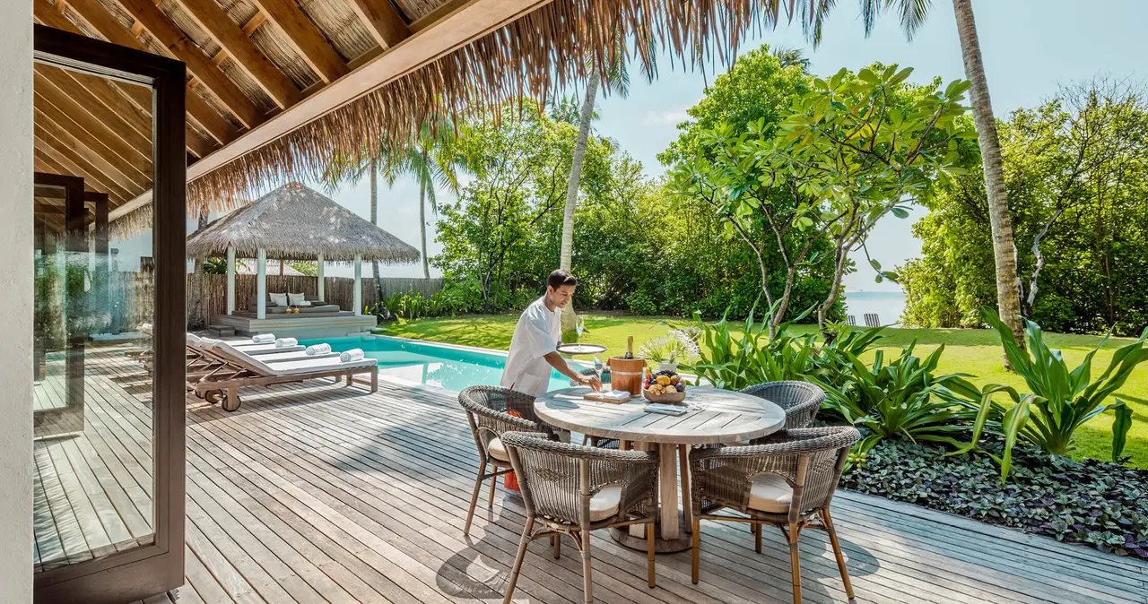 A table being laid on the deck of the Maalifushi Beach Villa, beside the pool and the garden.