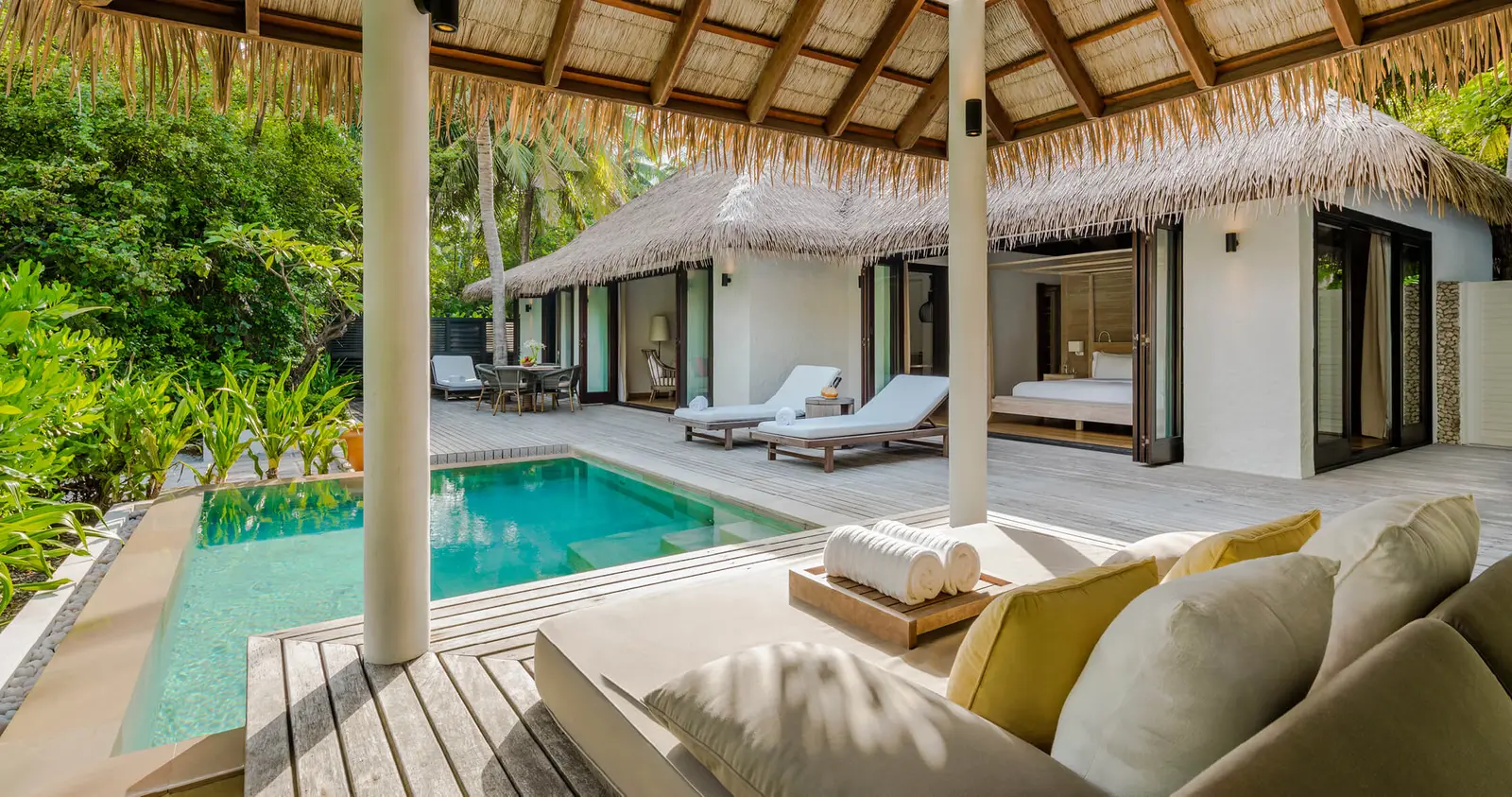 The covered deck of a Two-Bedroom Beach Villa, with the pool, loungers and yellow cushions.