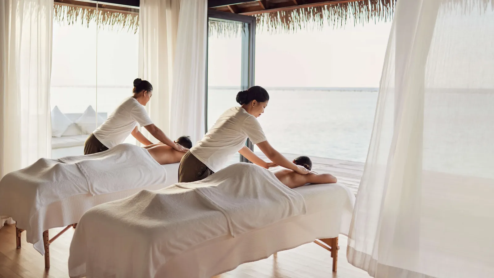 2 therapists giving a couple’s massage in an overwater spa room, its curtains open to the sea.