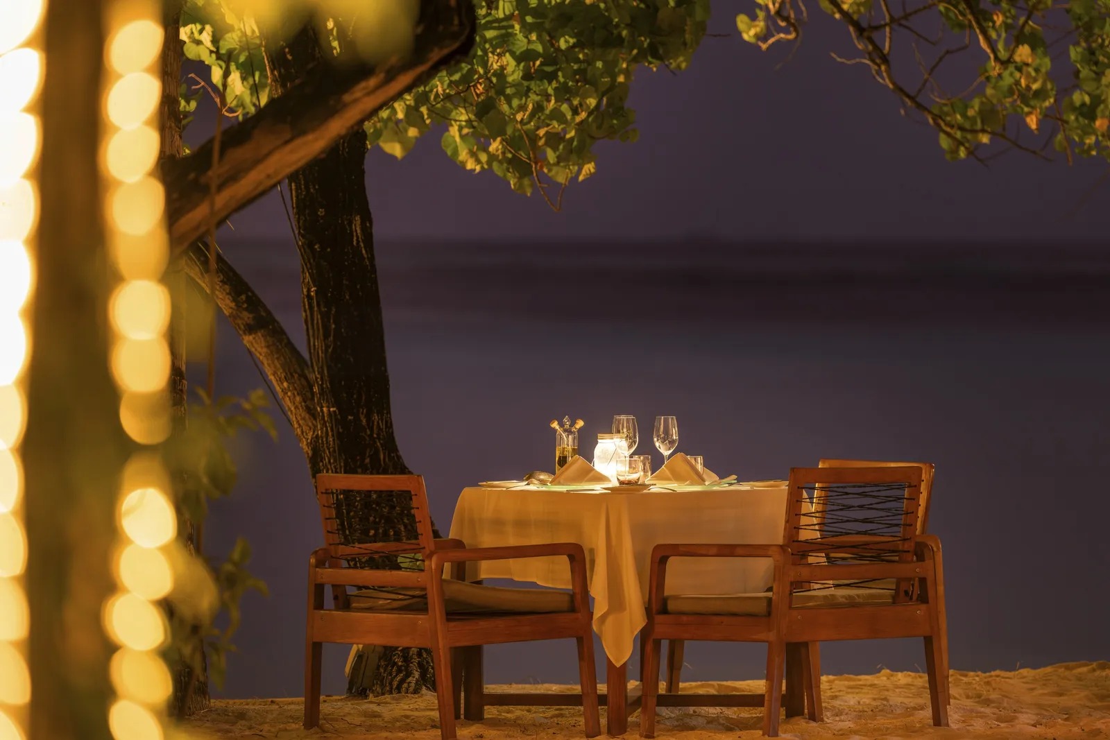 A table for 2 laid on the sand at Alizée at night, lights strung on a tree