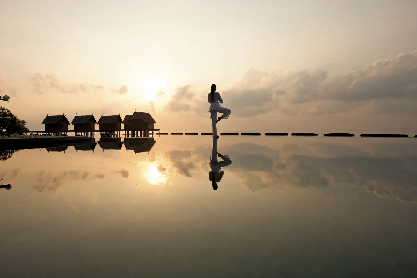 Someone holding a yoga pose at sunset, mirrored in the water, the water villas on the horizon