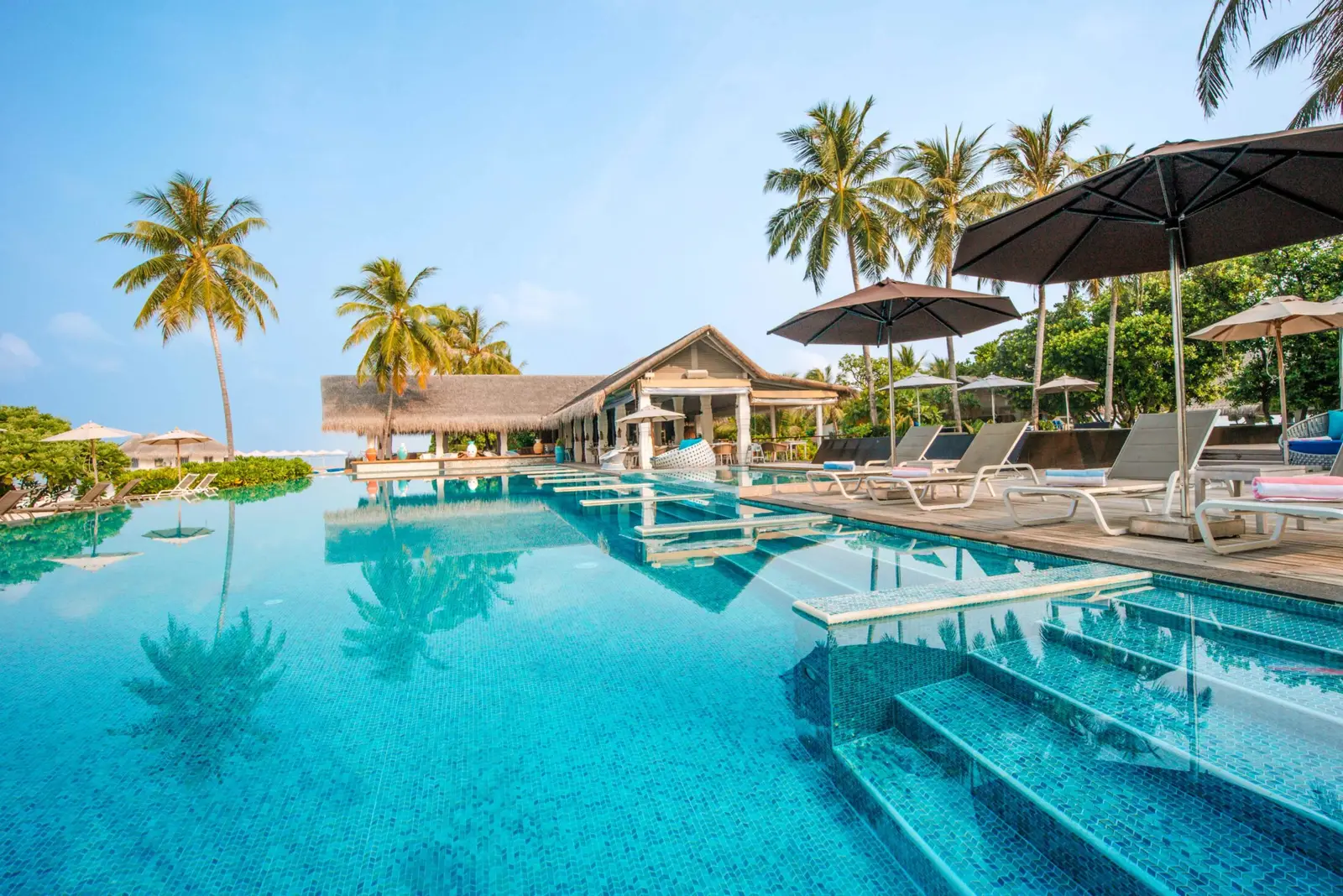 The main pool beside Acquapazza, with steps into the water, black parasols, palms and the pavilion at the far end.