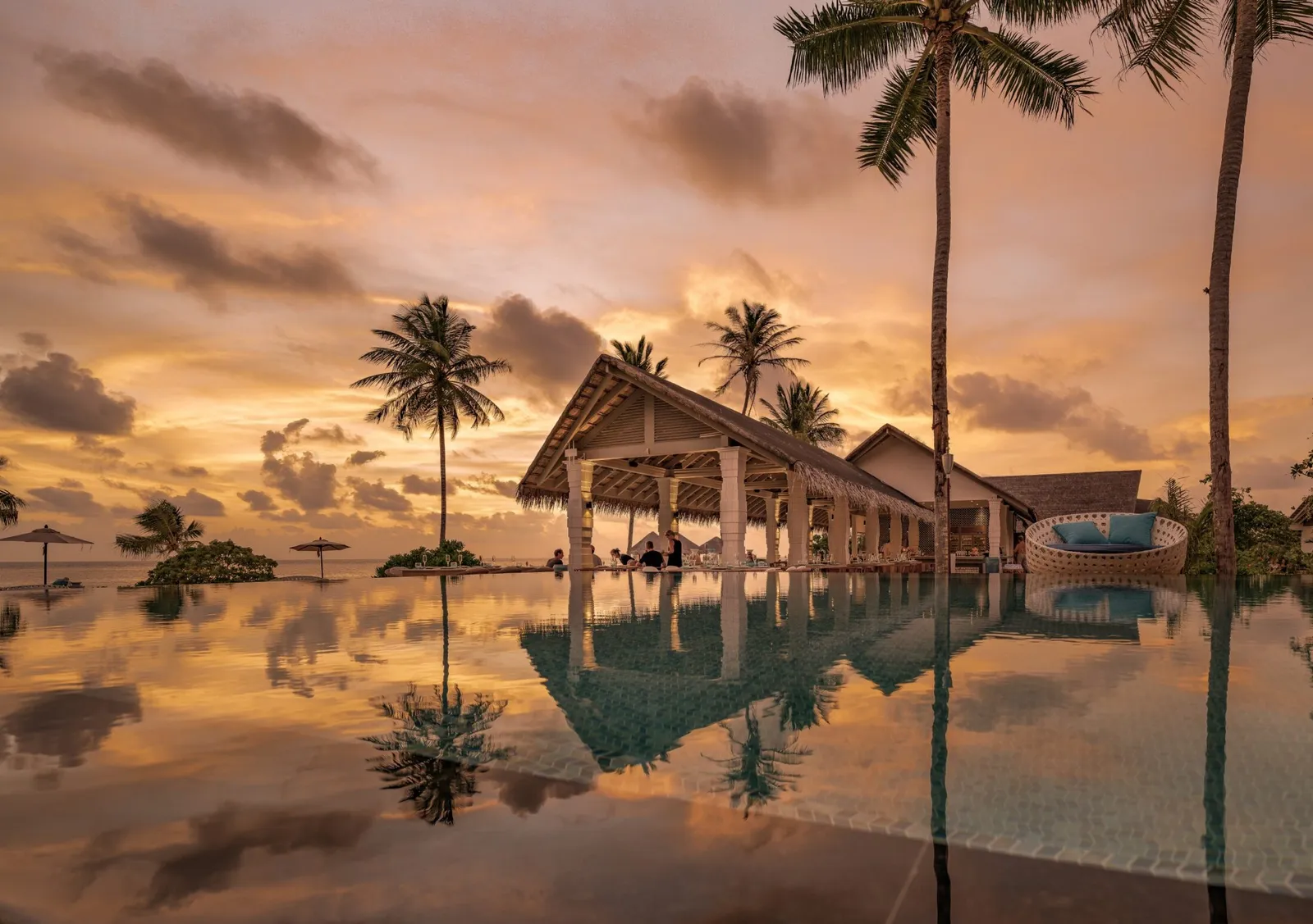 The pool at sunset: the orange sky and the palms mirrored in still water, in front of the thatched pavilion.