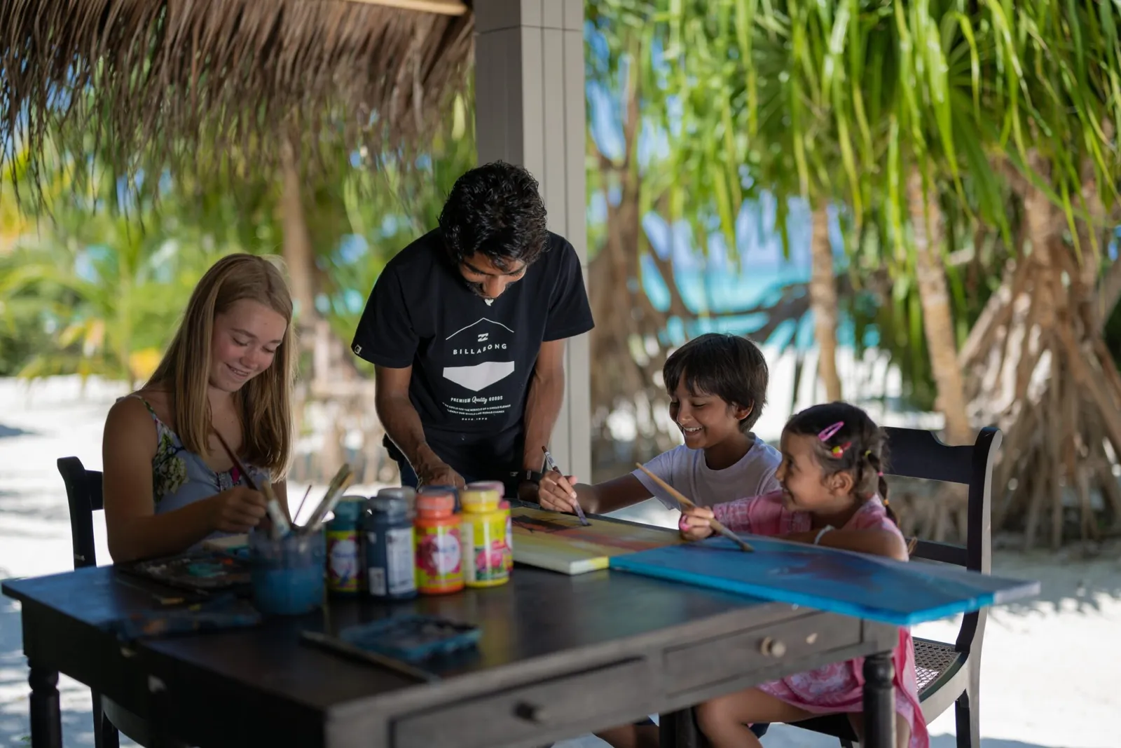 At the kids’ club 2 girls and a little girl paint at a table with a helper, under a thatched roof.