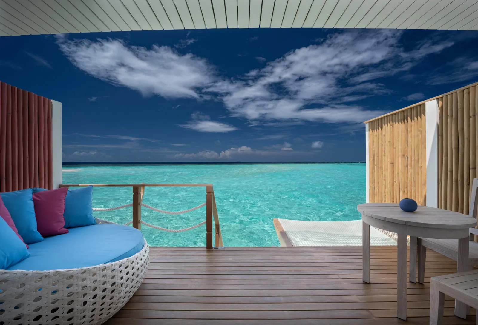 A Lagoon Villa deck over turquoise water, with the round daybed, the table and the steps down to the sea.