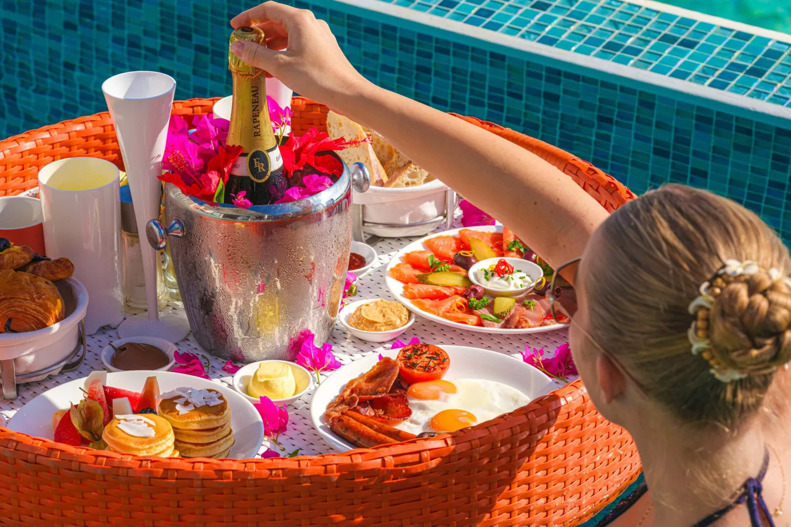 A woman breakfasts in her villa pool from a floating basket of fruit, pastries and a bottle on ice.