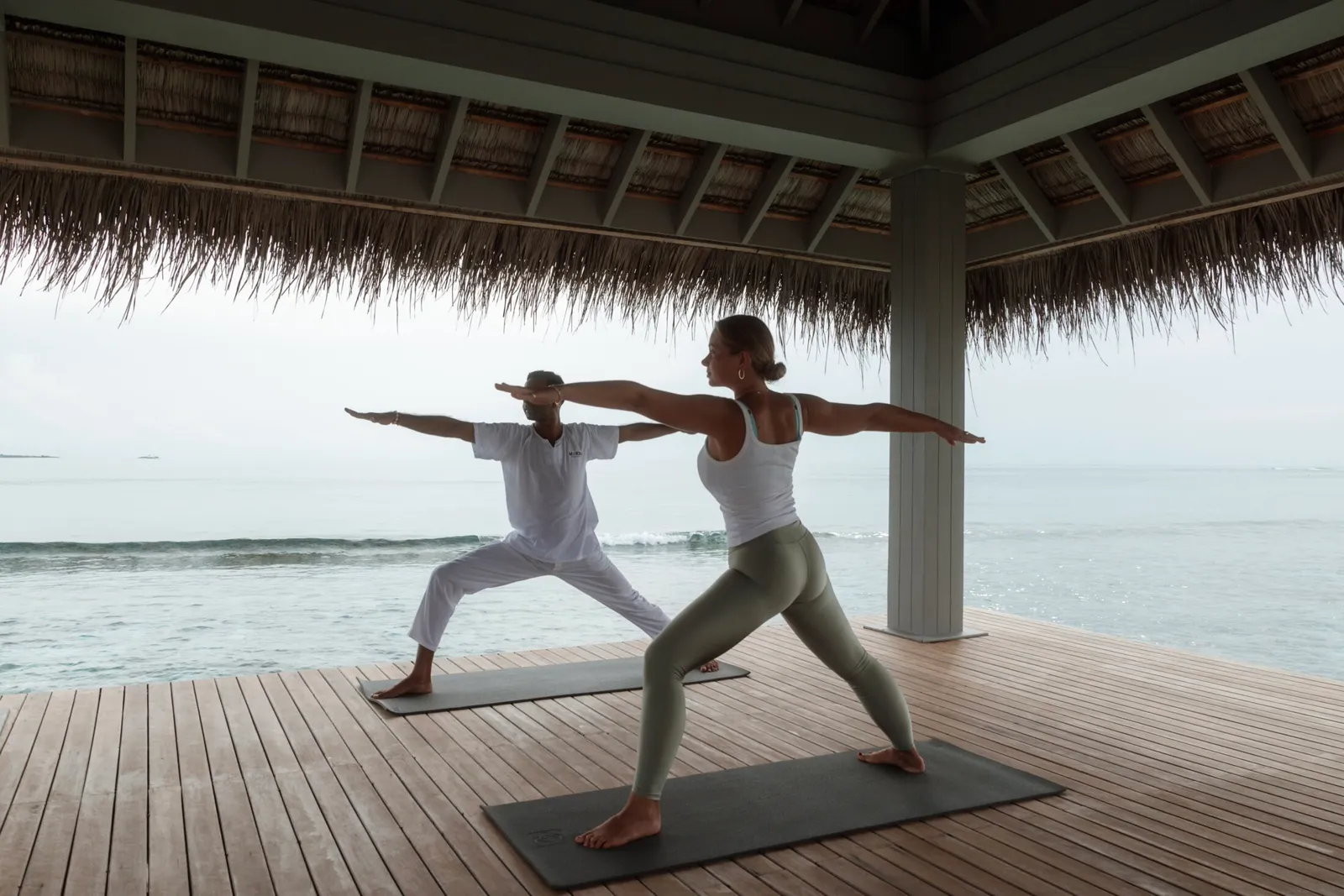 2 people hold a yoga pose in the thatched timber pavilion, facing the sea.