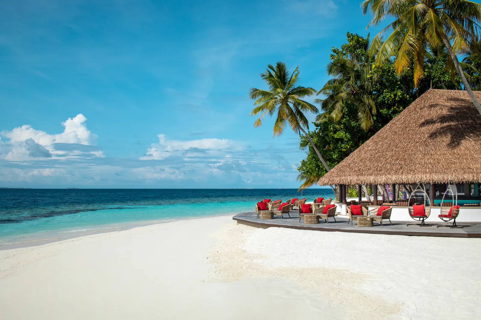 The beach bar under a great thatched roof, with woven armchairs and red cushions on the timber deck facing the sea.