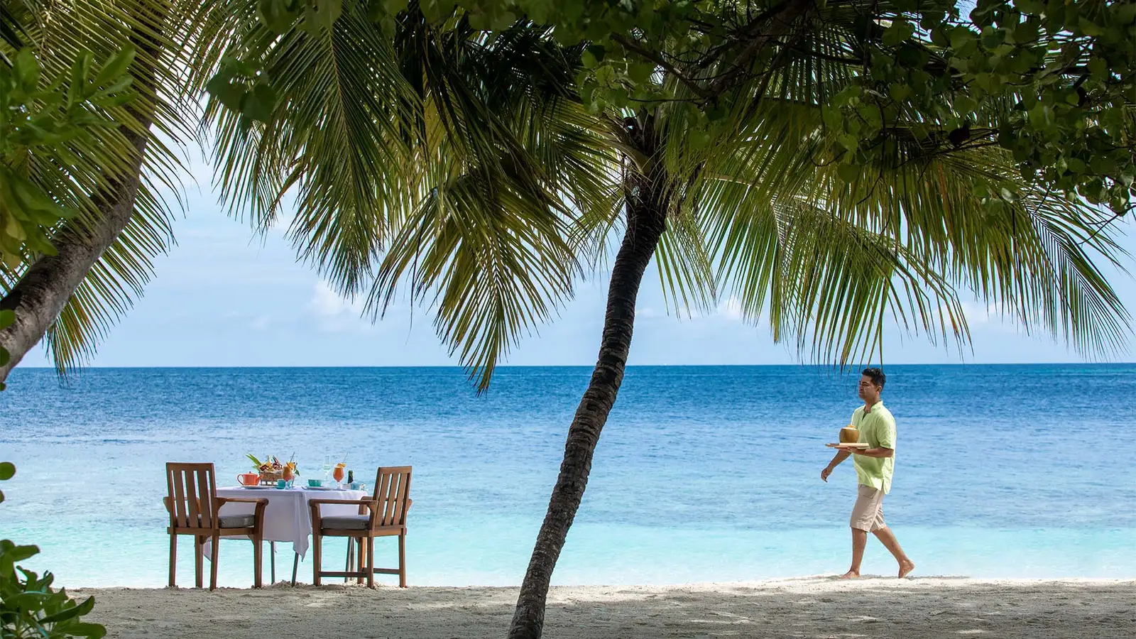 A table for 2 laid on the sand in the shade of a palm, and a waiter bringing breakfast.